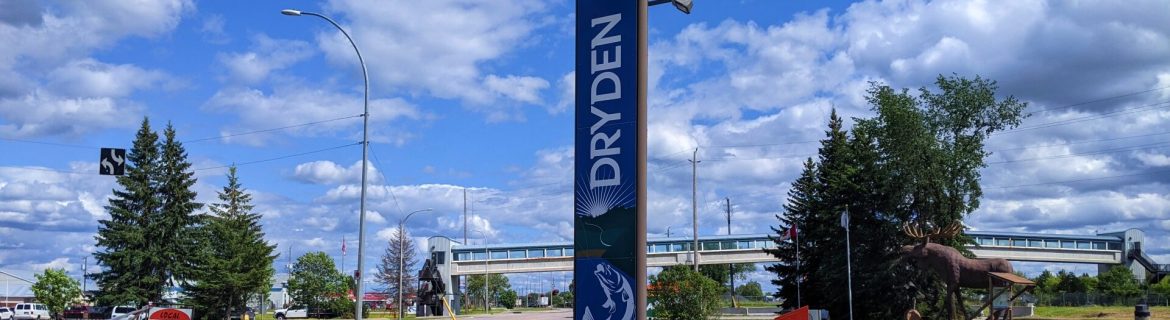 Tall, vertical Dryden sign with a silhouette of a fish on the bottom of the sign. Picnic bench in foreground. Trans Canada Highway to the left.